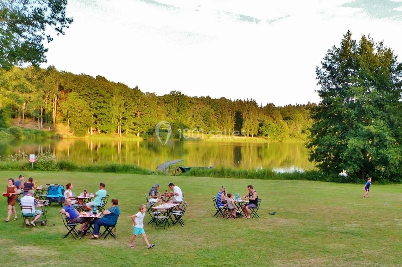 Des personnes dînent en plein air sur des tables installées près d'un lac entouré d'arbres.
