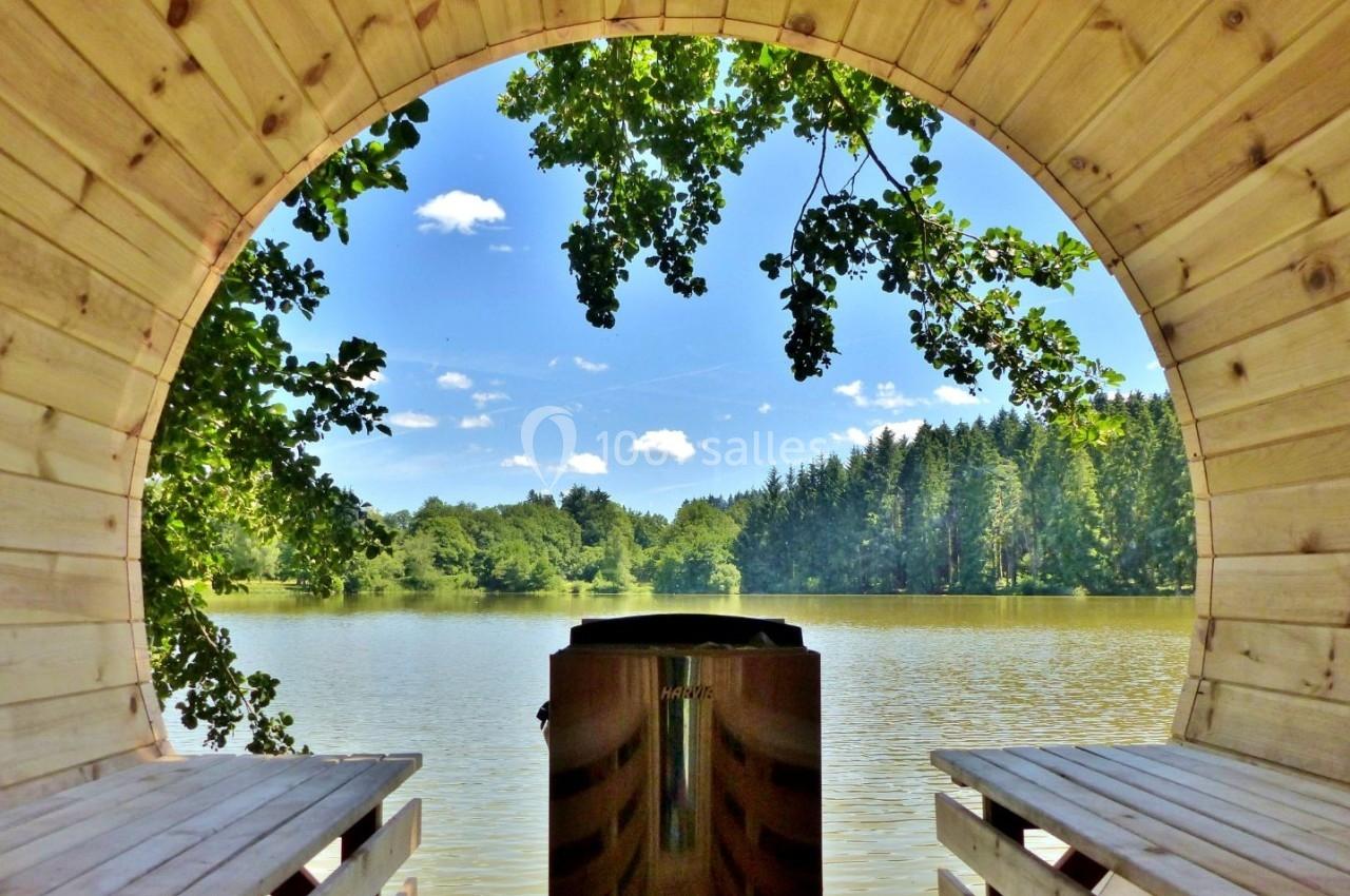 Vue d'un lac entouré de forêt à travers l'ouverture circulaire d'une cabane en bois avec un poêle au centre.