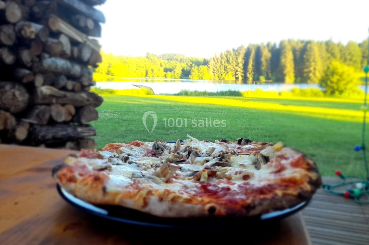 Pizza garnie de champignons et fromage sur une table en bois, avec vue sur un lac et une forêt en arrière-plan.