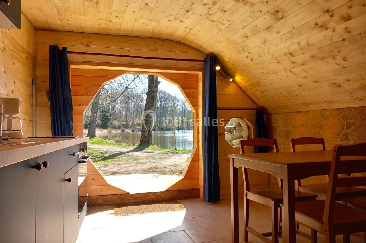 Vue intérieure d'une cabane en bois avec une grande ouverture circulaire donnant sur un paysage arboré et un étang.