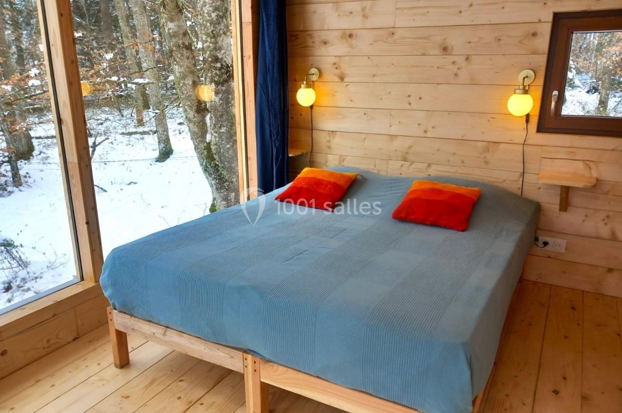 Lit double avec couverture bleue et coussins colorés dans une chambre en bois avec vue sur une forêt enneigée.