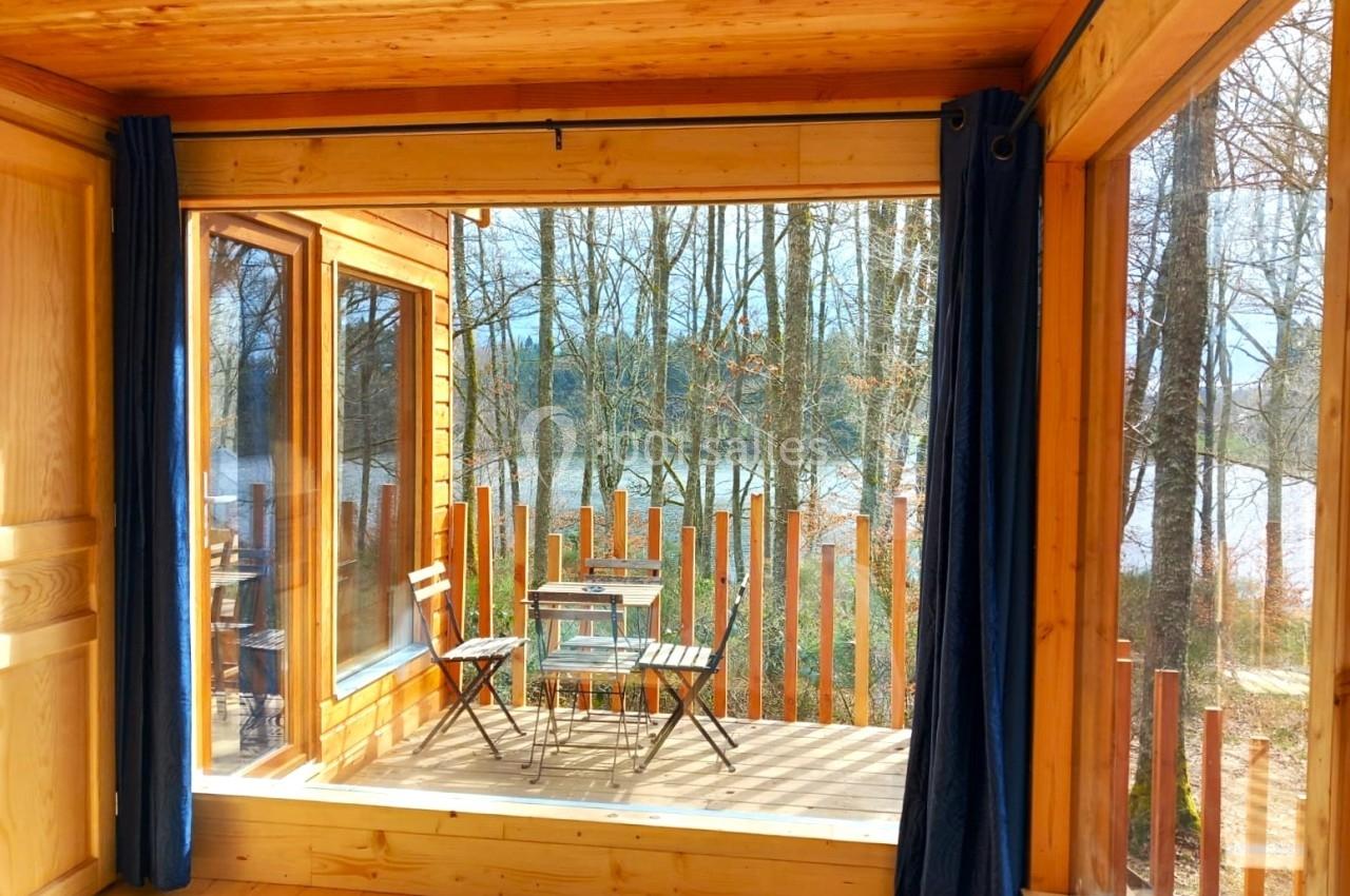 Vue depuis l'intérieur d'une cabane en bois sur une terrasse avec table, chaises et paysage boisé en arrière-plan.