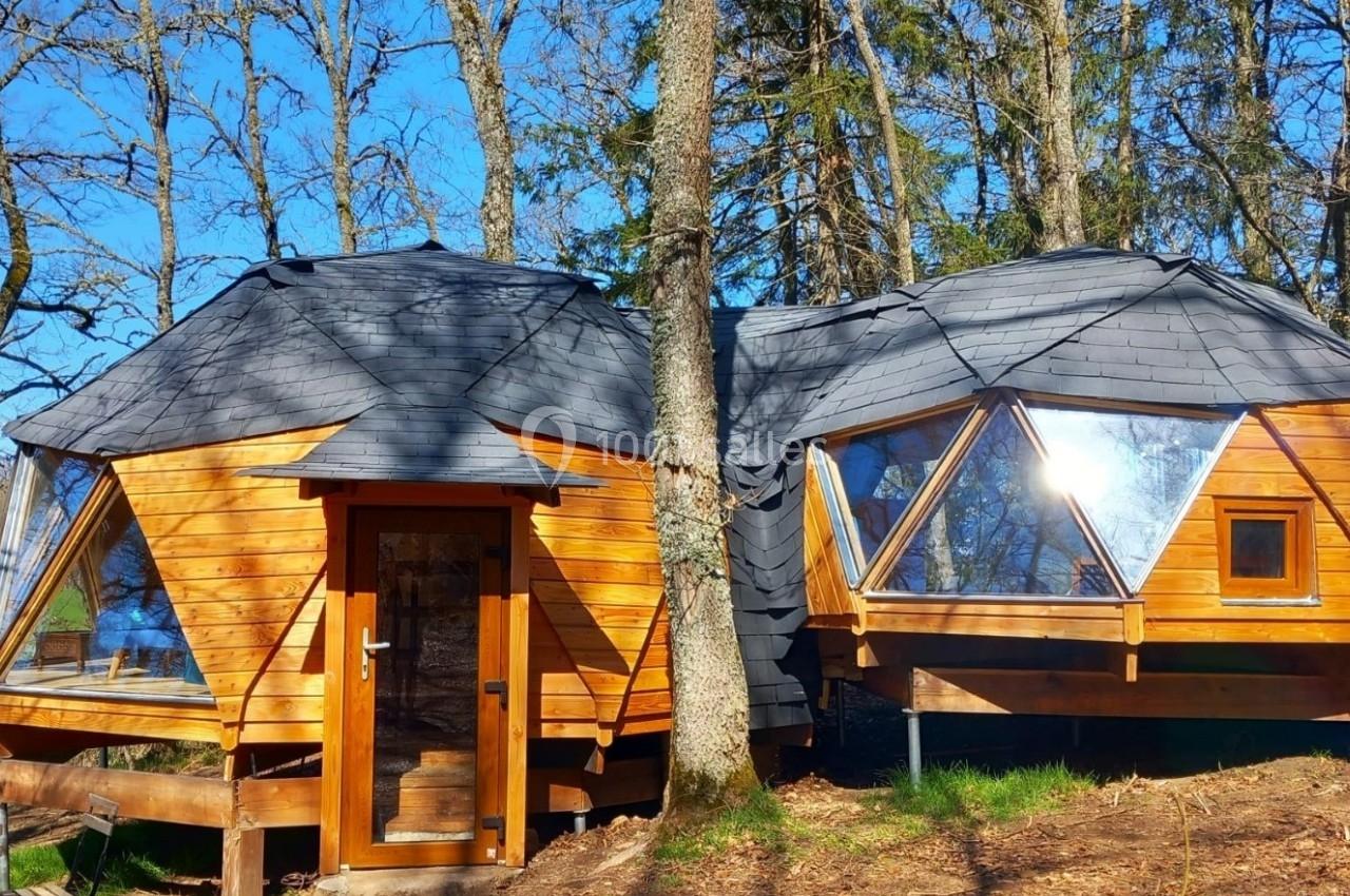 Cabane en bois avec des fenêtres géométriques, située dans une forêt ensoleillée.
