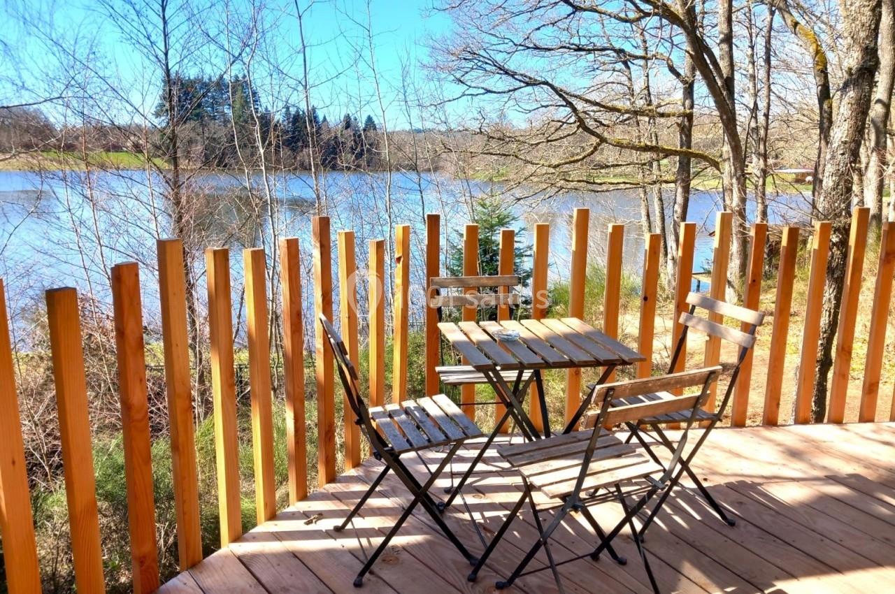 Terrasse en bois avec table et chaises pliantes, entourée d'une barrière, offrant une vue sur un lac et des arbres.
