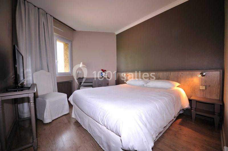 Chambre avec lit double, draps blancs, murs sombres, fenêtre avec rideaux, bureau et chaise blanche.