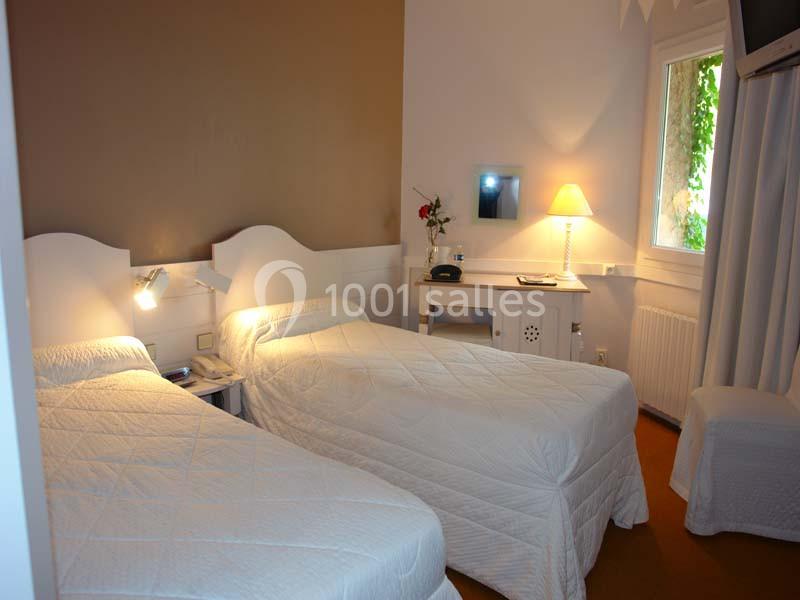 Deux lits simples avec couvre-lits blancs dans une chambre lumineuse, équipée d'un bureau, d'une lampe et d'une fenêtre.