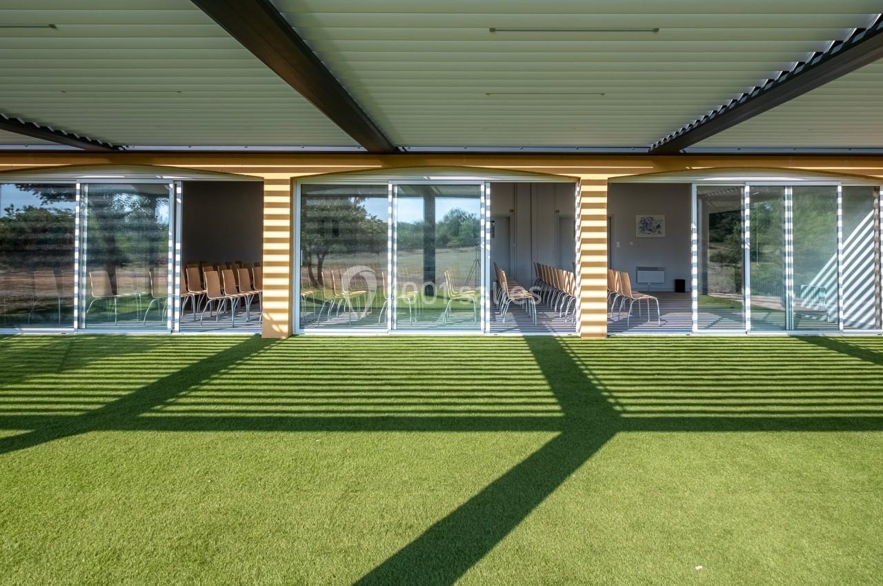Salle vitrée avec des chaises alignées à l'intérieur, vue depuis une terrasse couverte avec sol en gazon synthétique.
