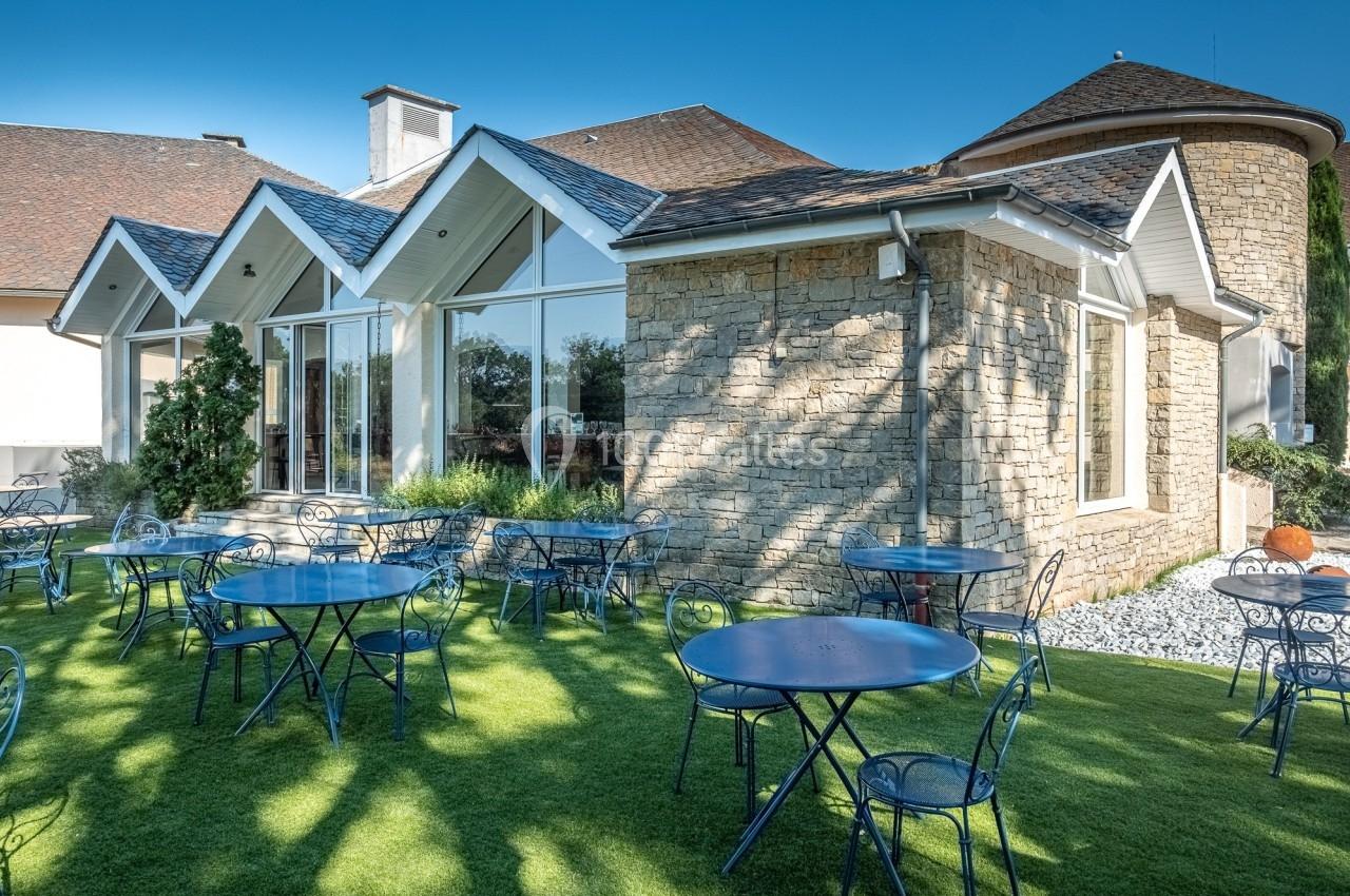 Terrasse avec tables et chaises en métal bleu sur une pelouse, devant un bâtiment en pierre avec de grandes fenêtres.