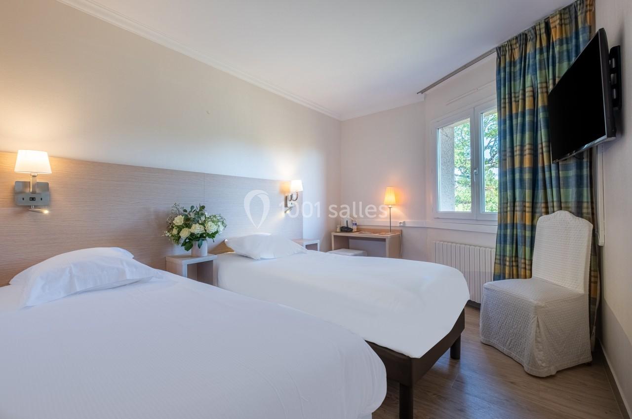 Chambre lumineuse avec deux lits simples, une chaise, une télévision murale et une fenêtre avec rideaux colorés.