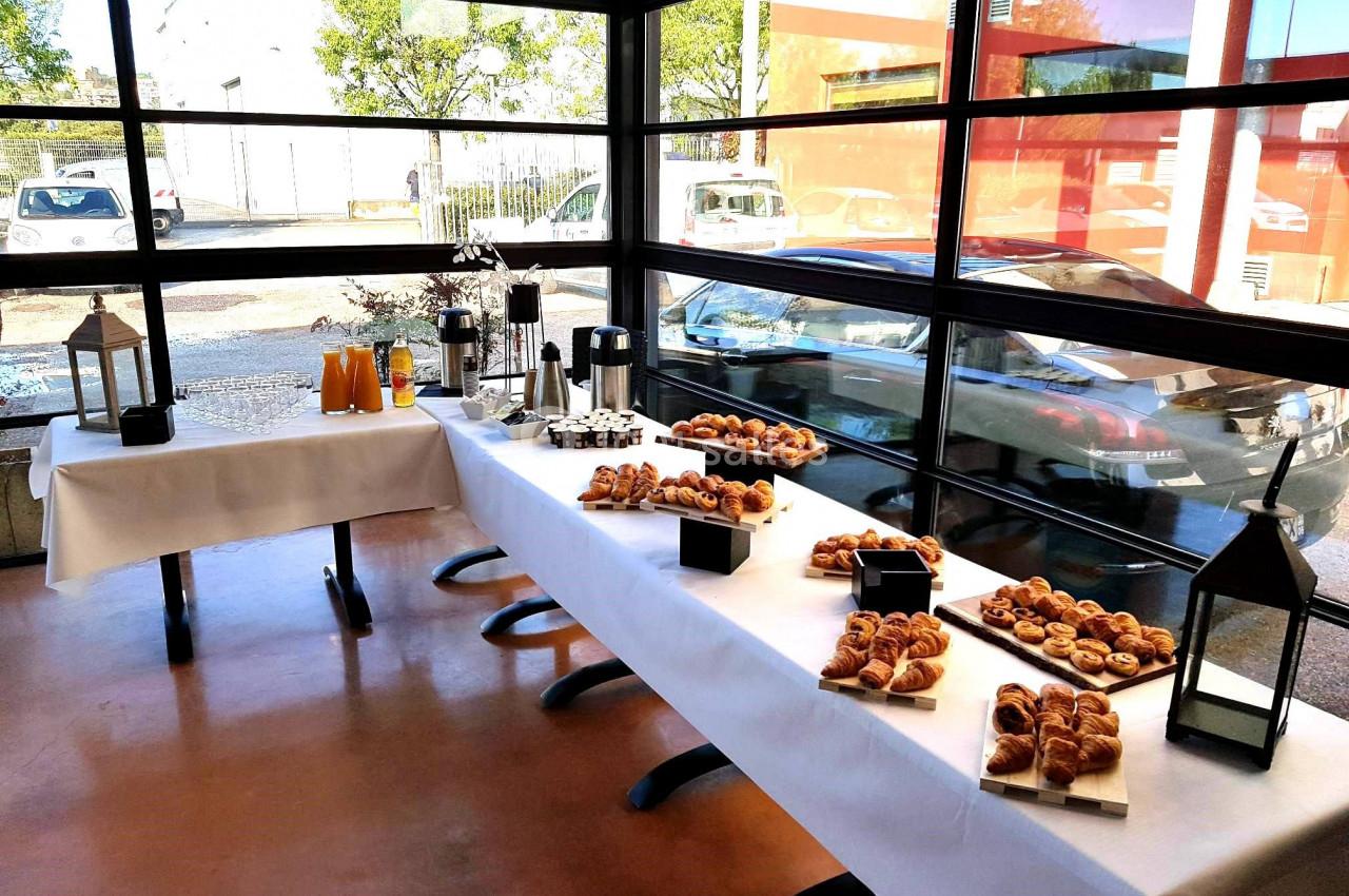 Buffet de viennoiseries, boissons et jus d'orange disposé sur des tables dans une salle lumineuse avec vue sur l'extérieur.