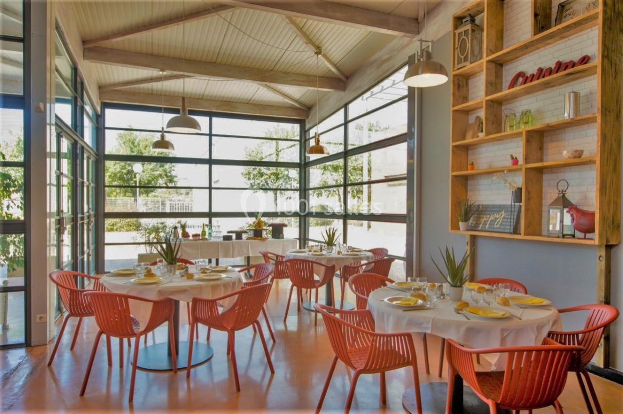 Salle lumineuse avec de grandes baies vitrées, tables dressées avec chaises rouges et étagères décoratives en bois.