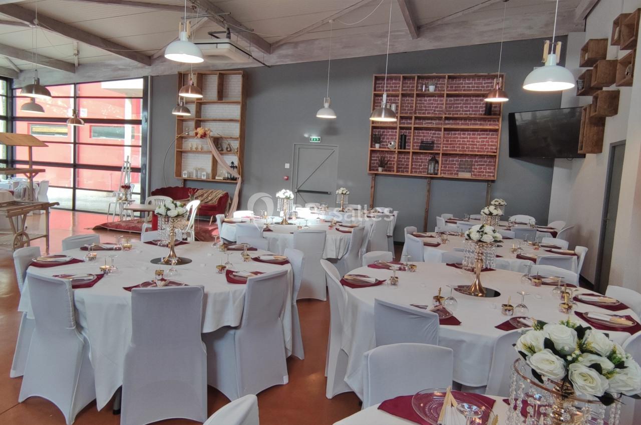 Salle de réception lumineuse avec tables rondes dressées, nappes blanches, décorations florales et étagères en arrière-plan.