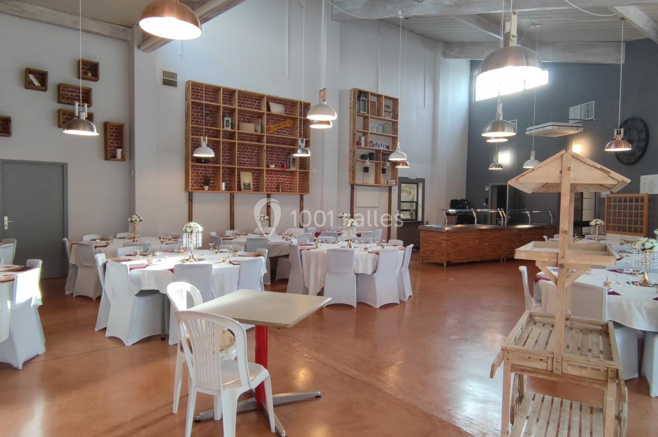 Salle de réception lumineuse avec tables rondes dressées, chaises blanches et décorations florales.