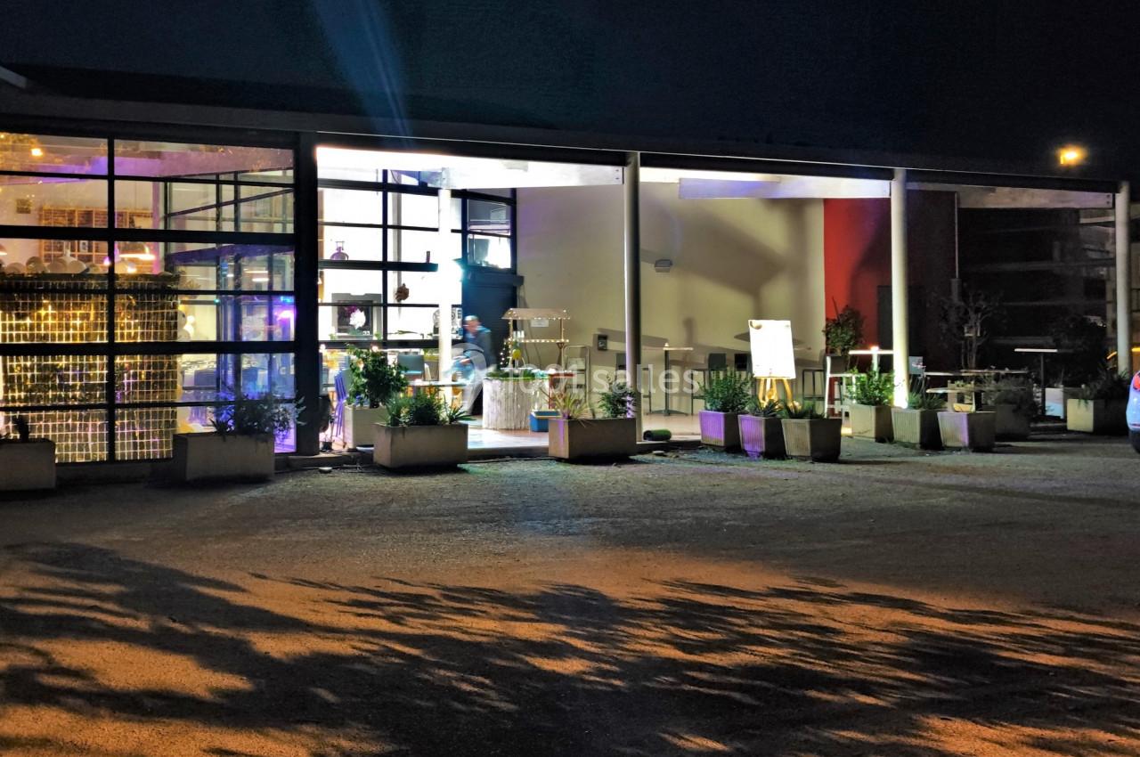 Façade vitrée d'un bâtiment moderne éclairé la nuit, entourée de plantes en pots et d'un parking.