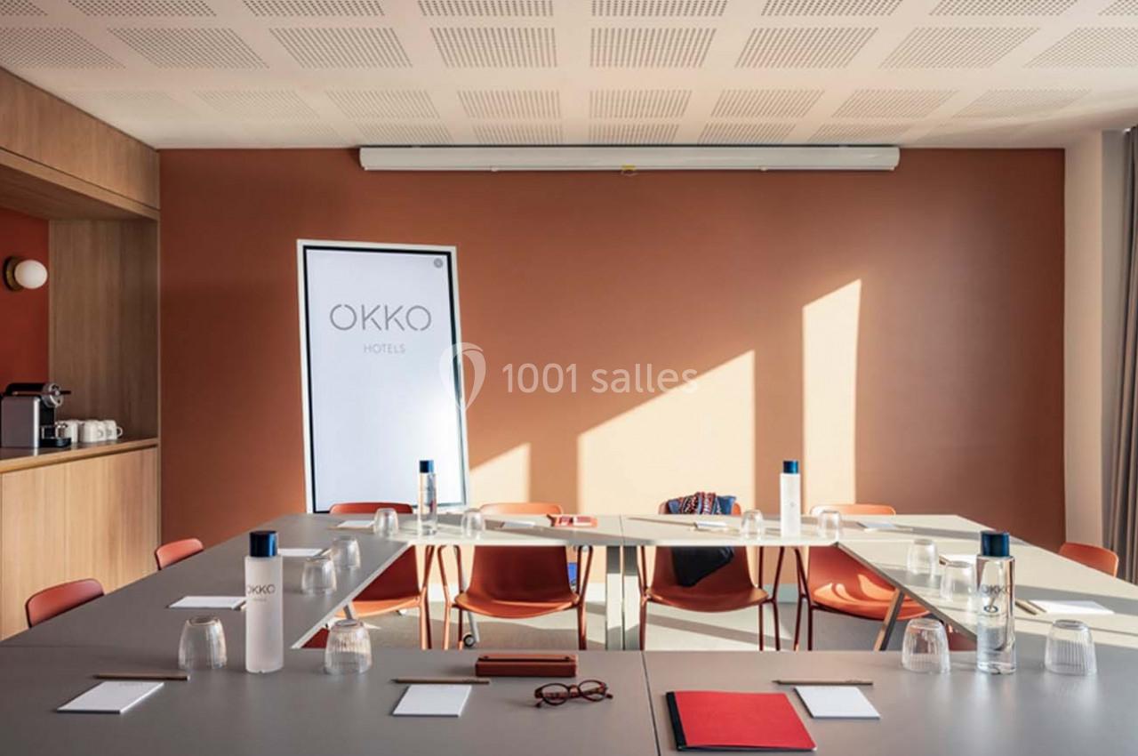 Salle de réunion lumineuse avec table en U, chaises rouges, bouteilles d'eau, verres et écran affichant ’OKKO’.