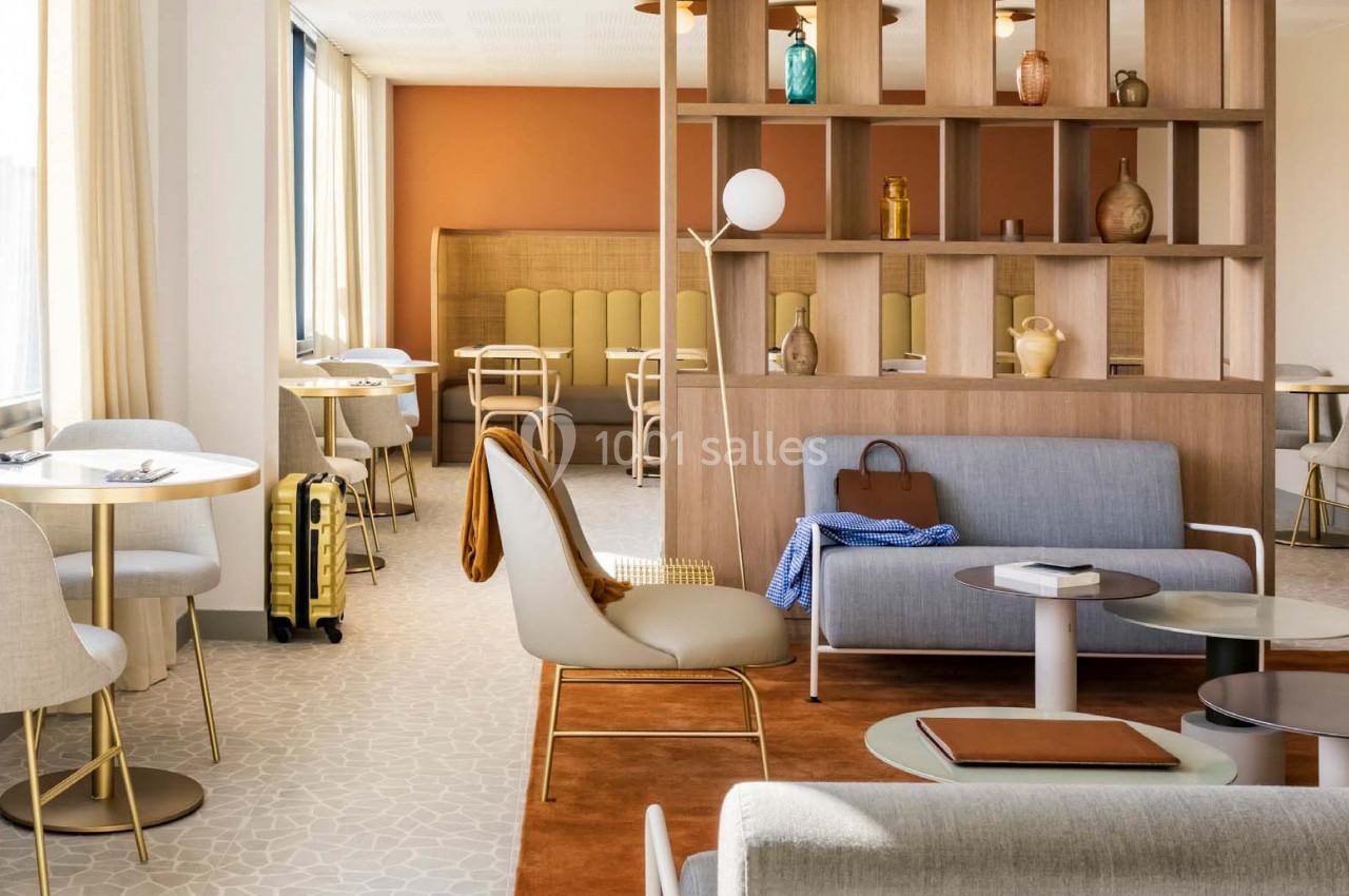 Espace salon lumineux avec mobilier moderne, cloison en bois, tables, chaises et valise jaune près d'une fenêtre.