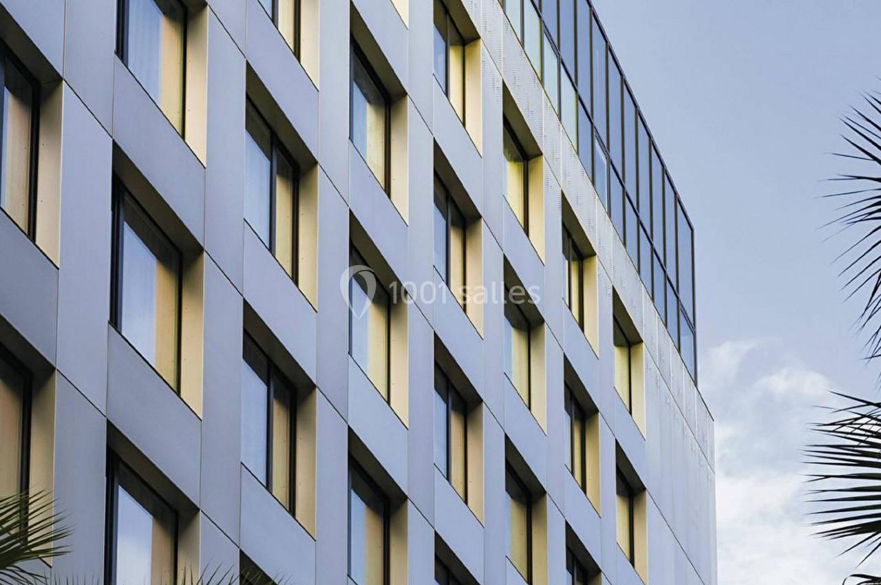 Façade d'un immeuble moderne avec des fenêtres alignées, entourée de palmiers sous un ciel partiellement dégagé.
