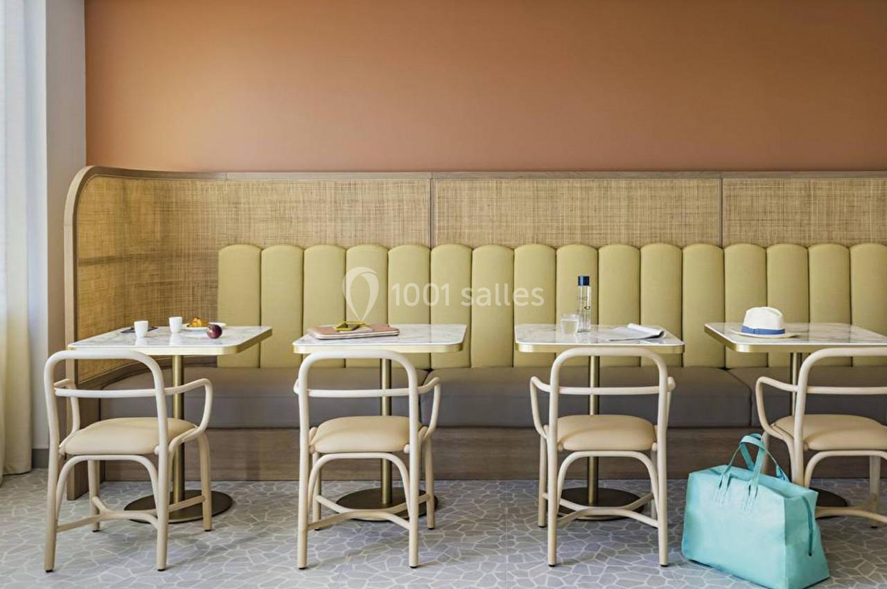 Salle de restaurant avec banquette jaune, tables en marbre, chaises en bois clair et sac turquoise posé au sol.