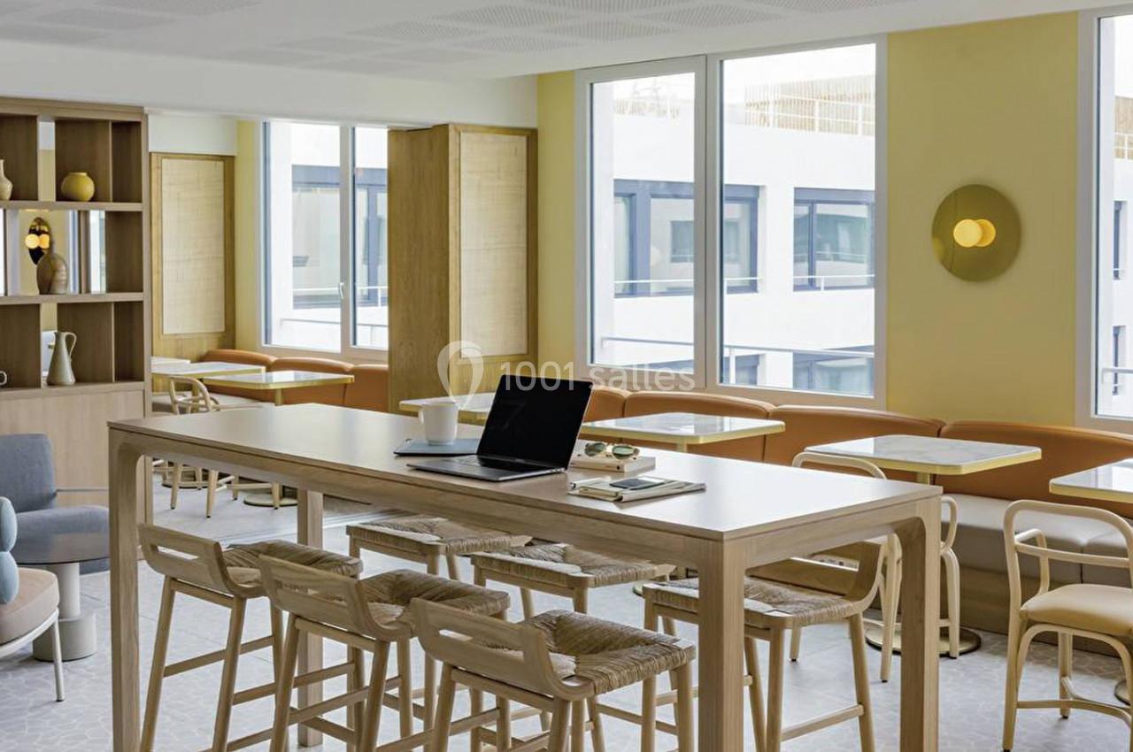 Espace lumineux avec tables, chaises et étagères, décoré dans des tons clairs, avec un ordinateur posé sur une table haute.