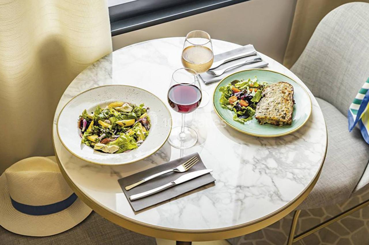 Table ronde en marbre avec deux assiettes de plats, deux verres de vin, des couverts et un chapeau posé sur une chaise.