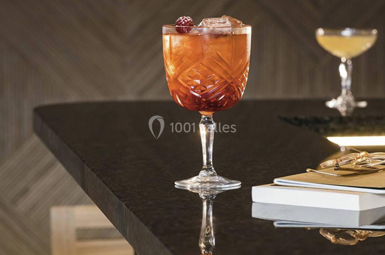 Verre à cocktail rouge garni de fruits rouges et glaçons, posé sur un comptoir sombre avec des carnets à proximité.