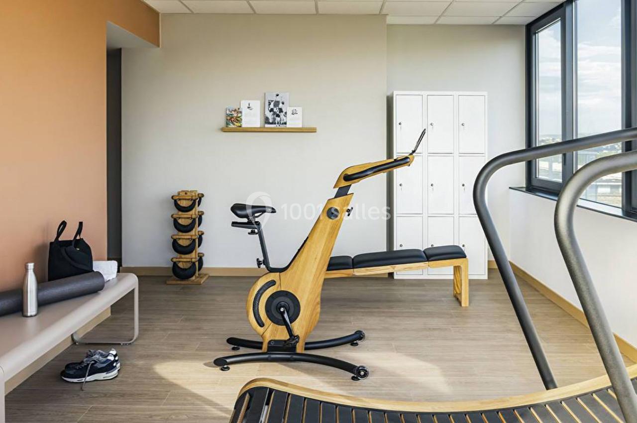 Salle de sport lumineuse avec vélo d'exercice en bois, haltères, banc de musculation et casiers blancs.