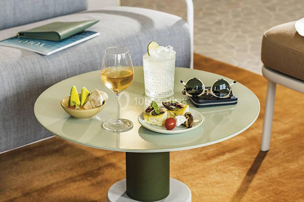 Table basse avec verre de vin, cocktail, assiette de tapas, lunettes de soleil et livre dans un salon lumineux.