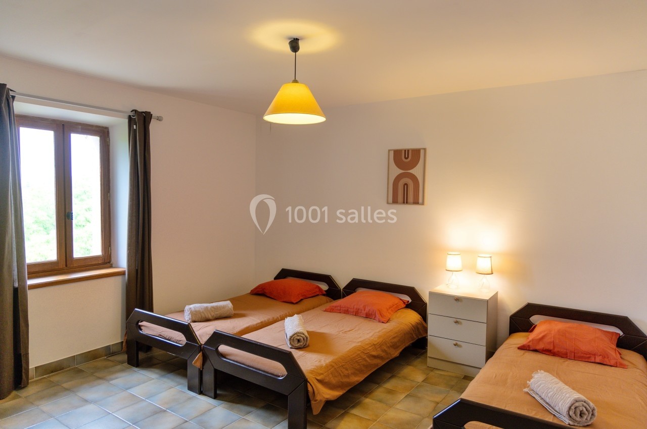 Chambre avec trois lits simples, draps orange, serviettes pliées, lampe suspendue et fenêtre avec rideaux.