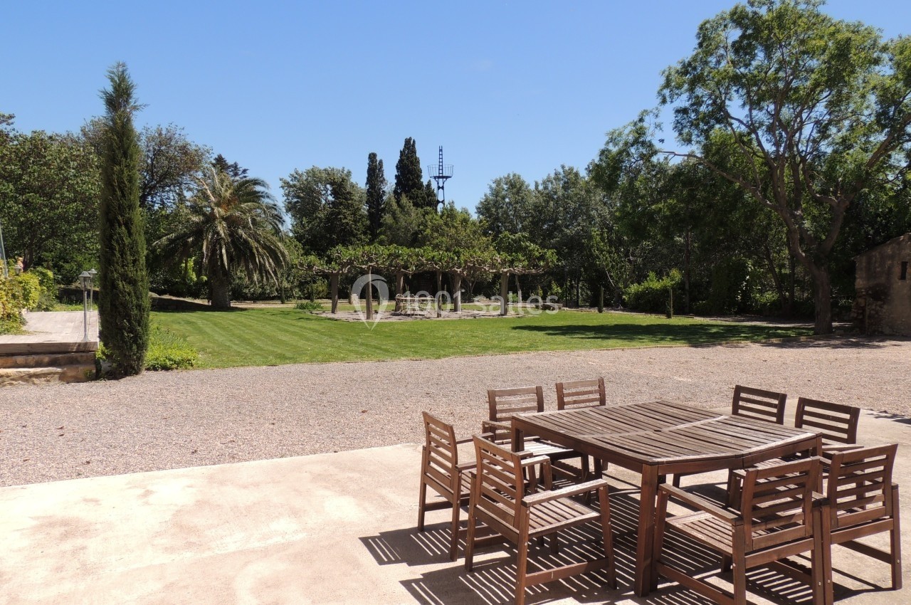 Table et chaises en bois sur une terrasse, avec un jardin arboré et une pelouse en arrière-plan sous un ciel dégagé.