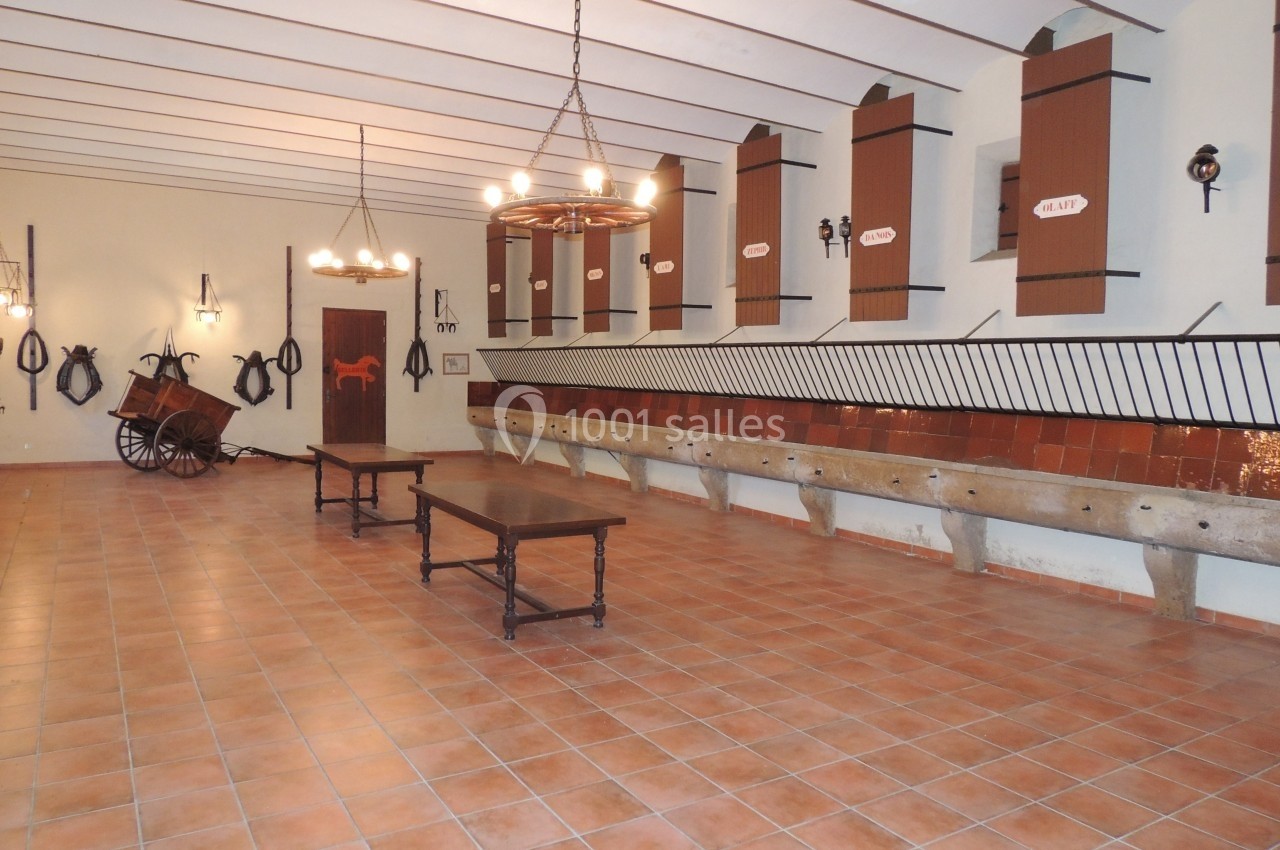 Salle avec mangeoires en pierre, tables en bois, murs décorés d'outils équestres et plafonniers suspendus.