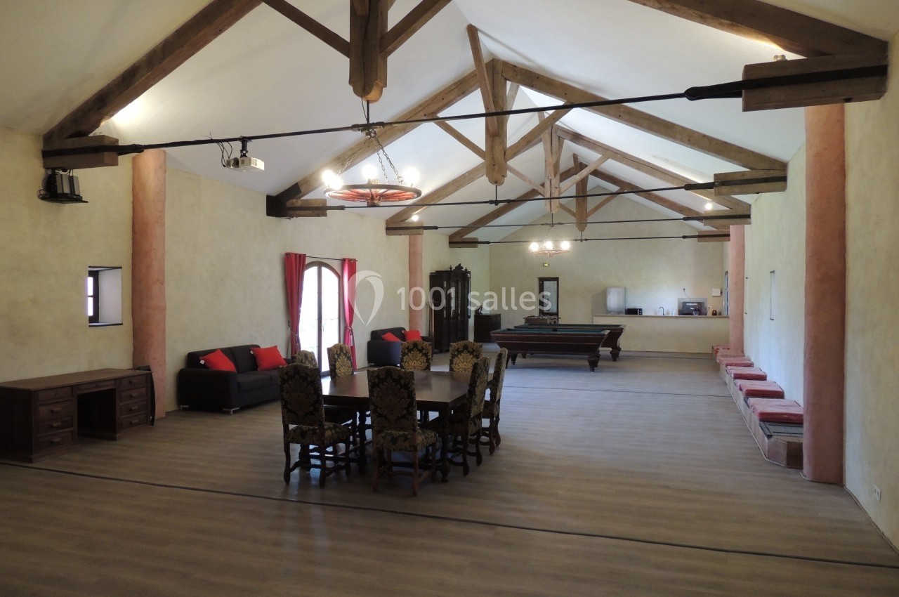 Salle spacieuse avec poutres apparentes, table et chaises au centre, canapés et billard en arrière-plan.