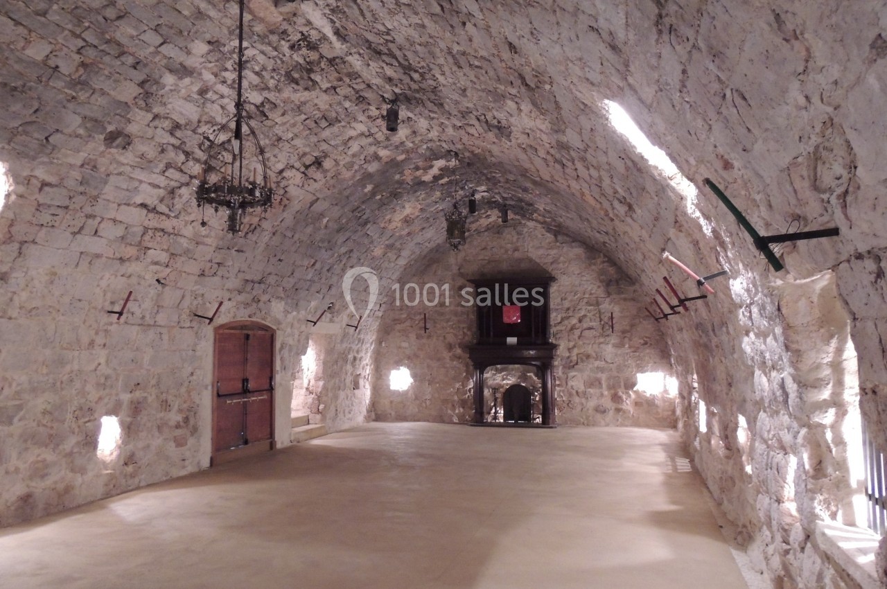 Salle voûtée en pierre avec un sol en béton, des chandeliers suspendus et une cheminée au fond.