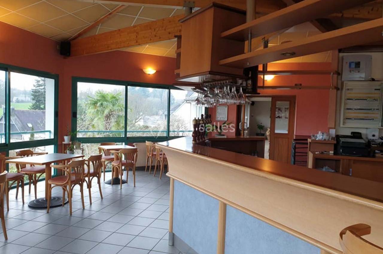 Salle de restaurant lumineuse avec grandes baies vitrées, mobilier en bois et comptoir de bar en premier plan.