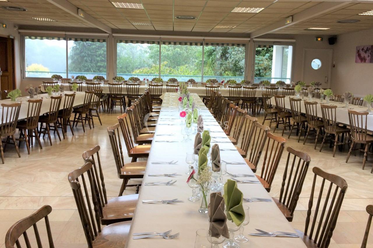 Salle de réception avec de longues tables dressées, nappes blanches, serviettes pliées et chaises en bois.