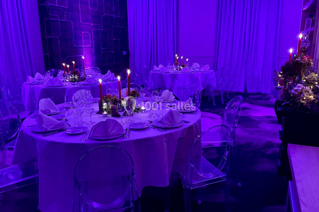 Salle de réception éclairée en violet avec tables rondes dressées, nappes blanches, bougies et décorations florales.