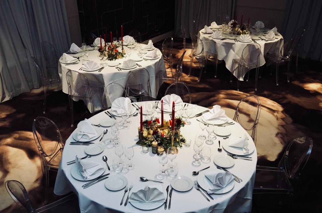 Tables rondes élégamment dressées avec nappes blanches, chandeliers rouges et décorations florales dans une salle feutrée.