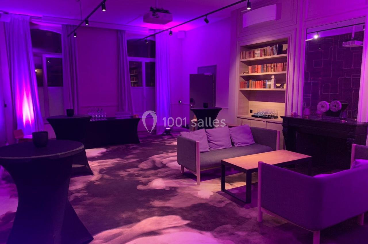 Salle de réception éclairée de lumières violettes, avec canapés, tables basses, bibliothèques et nappes noires.