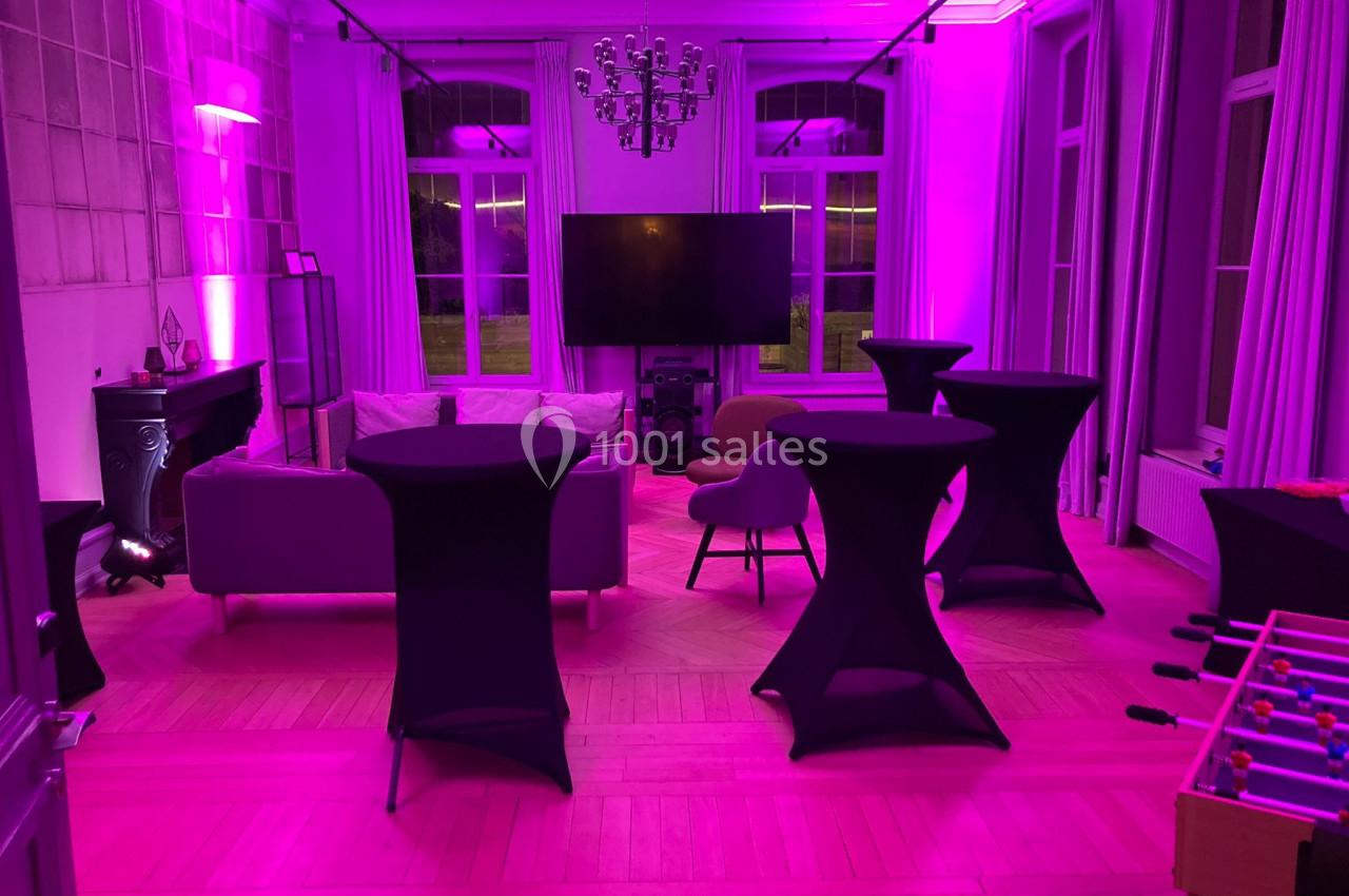 Salle lumineuse avec éclairage rose, tables hautes noires, canapé, télévision murale et baby-foot au premier plan.