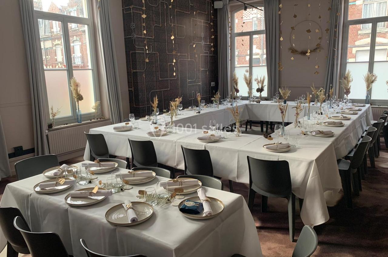 Salle de réception lumineuse avec tables dressées en U, nappes blanches, vaisselle élégante et décorations florales.
