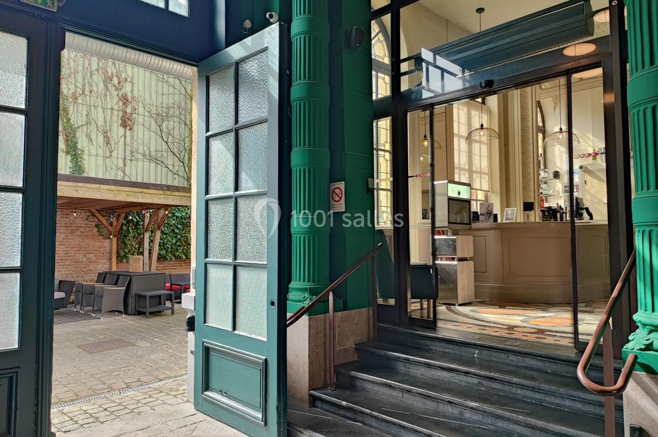 Entrée d'un bâtiment avec des portes ouvertes, escalier menant à un intérieur lumineux et terrasse extérieure aménagée.