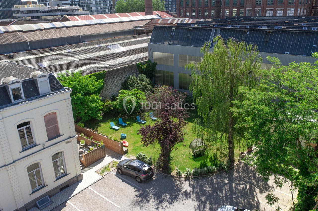 Vue aérienne d'une cour arborée avec pelouse, chaises longues, arbres et bâtiments industriels en arrière-plan.