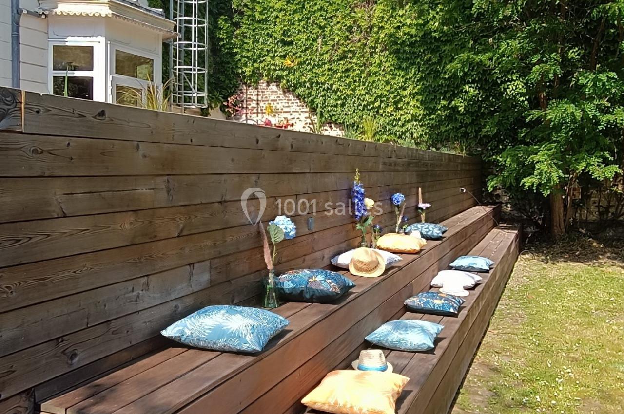 Bancs en bois avec coussins colorés disposés en terrasse dans un jardin ensoleillé, bordé d'arbres et d'une maison.