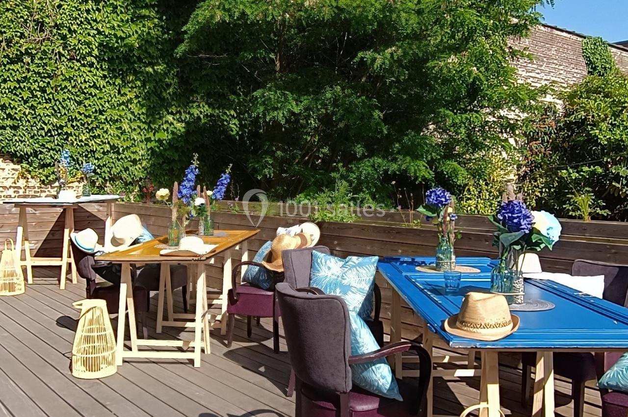 Terrasse en bois aménagée avec des tables, chaises et décorations, entourée de verdure sous un ciel bleu.