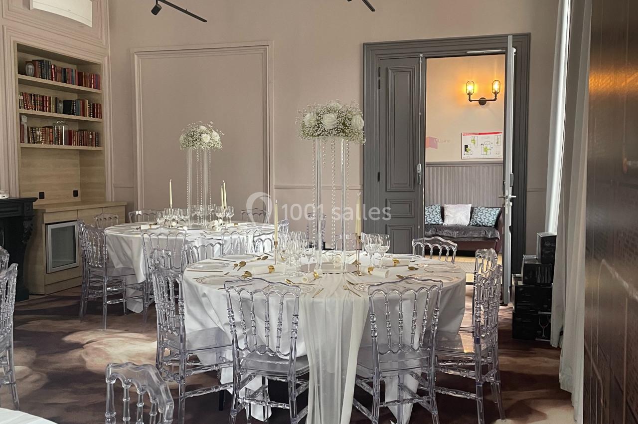 Salle élégante décorée pour un événement, avec tables rondes, nappes blanches, chaises transparentes et centres de table…