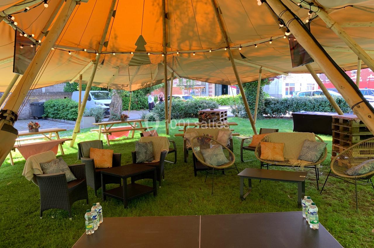 Espace extérieur aménagé sous une tente avec tables, fauteuils, guirlandes lumineuses et verdure en arrière-plan.