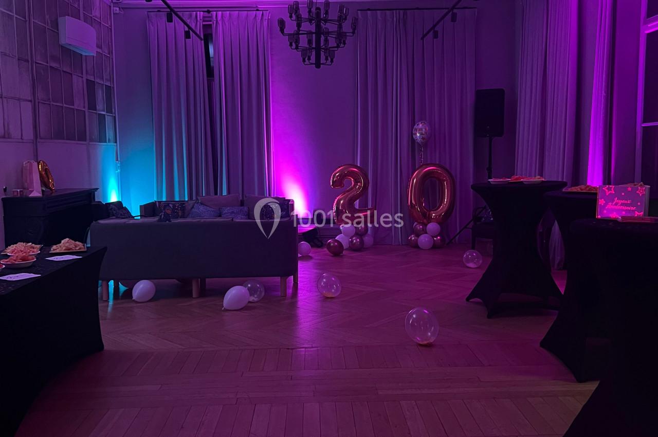 Salle décorée pour une fête avec éclairage violet, ballons ’20’, tables hautes et canapé en arrière-plan.