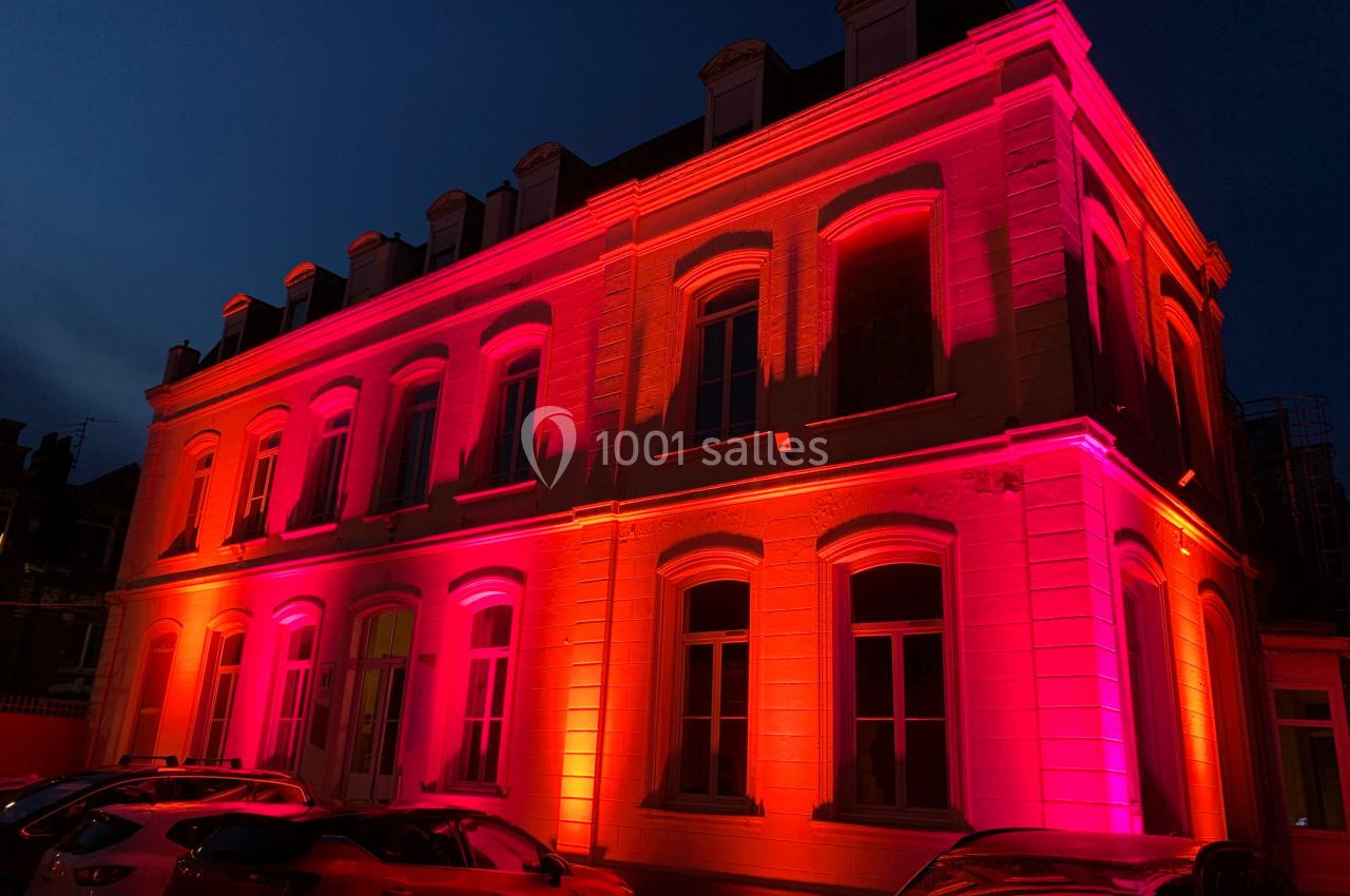 Façade d'un bâtiment éclairée par des lumières roses et oranges, avec des voitures garées au premier plan.