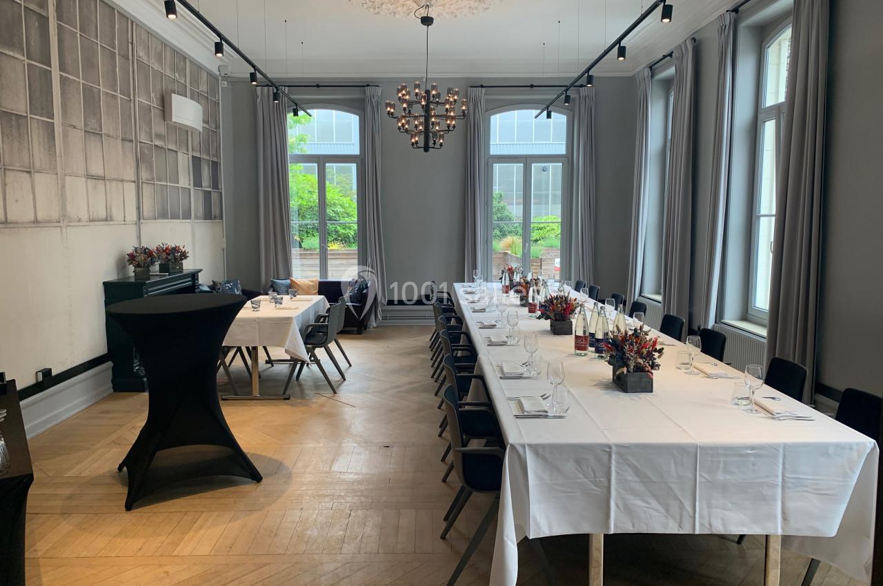 Salle lumineuse avec de grandes fenêtres, tables dressées avec nappes blanches, chaises noires et décorations florales.