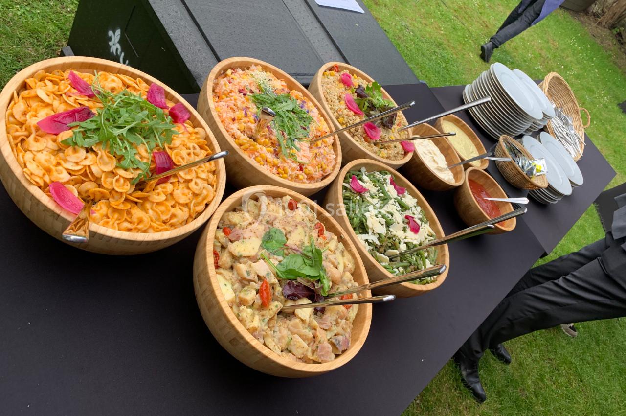 Buffet en plein air avec des salades variées présentées dans des bols en bois, accompagné d'assiettes et de couverts.