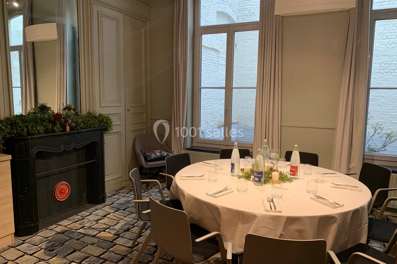 Salle lumineuse avec table ronde dressée, chaises modernes, cheminée décorée et grandes fenêtres donnant sur une cour.